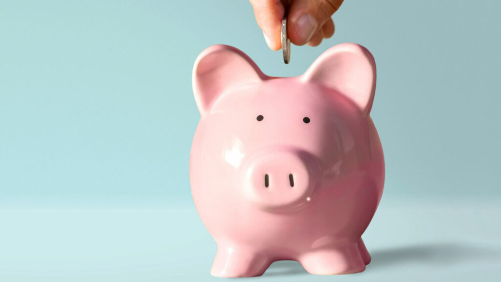 Fingers placing a coin into a piggy bank on a light blue background